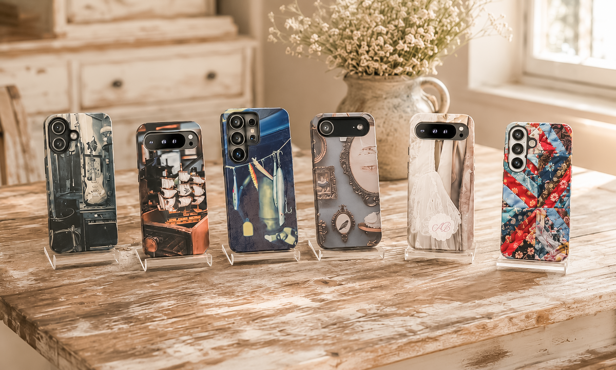 Six Vintage Hearts Club phone cases displayed on clear acrylic stands on a rustic farmhouse table, featuring vintage guitar, pirate ship, fishing lure, blue oval frame, bridal lace monogram, and colorful quilt designs.